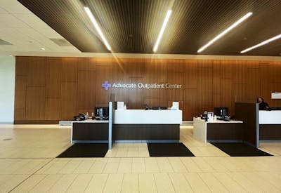 The main reception lobby of the Advocate Outpatient Center. A long wood-paneled feature wall carries the facility’s name and purple cross logo.